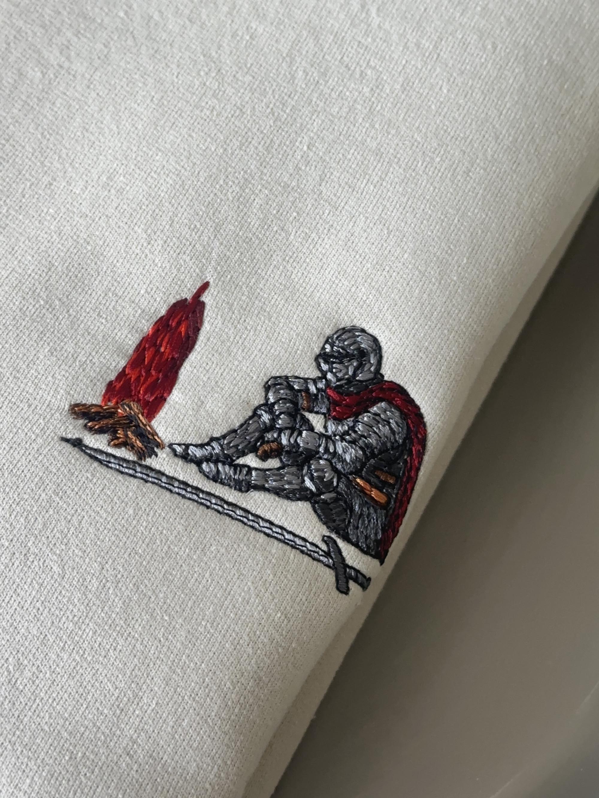 Close-up of a white or off-white fabric item featuring an embroidered design of a kneeling armored knight with a red cape next to a red and orange flame.