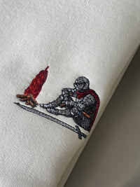 Close-up of a white or off-white fabric item featuring an embroidered design of a kneeling armored knight with a red cape next to a red and orange flame.